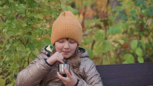 Kid Blows on Warm Drink and Tests Temperature with Finger in Autumn Park
