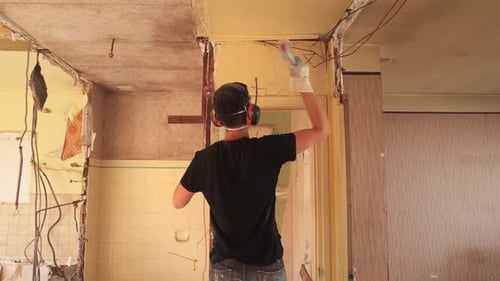 Man breaking wall and door frame inside demolition apartment with sledge hammer, static wide shot