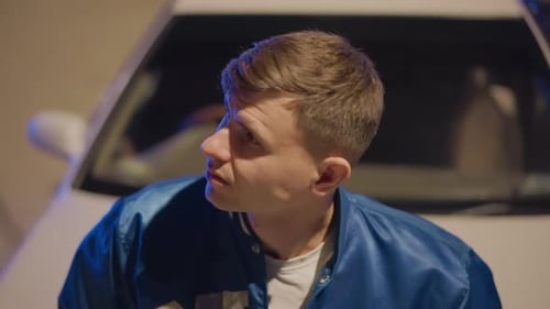 Young Man Leans Against Car, Looks Around At Night