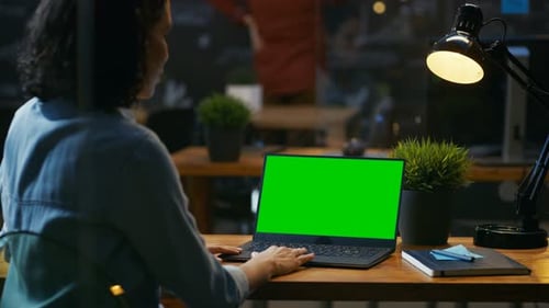 Female Office Worker at Her Desk Works on a Laptop with Mock-up Green Screen. Over the Shoulder Foo