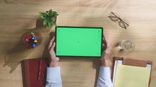 Top down view of man's hands holding a green screen tablet on a wooden desk
