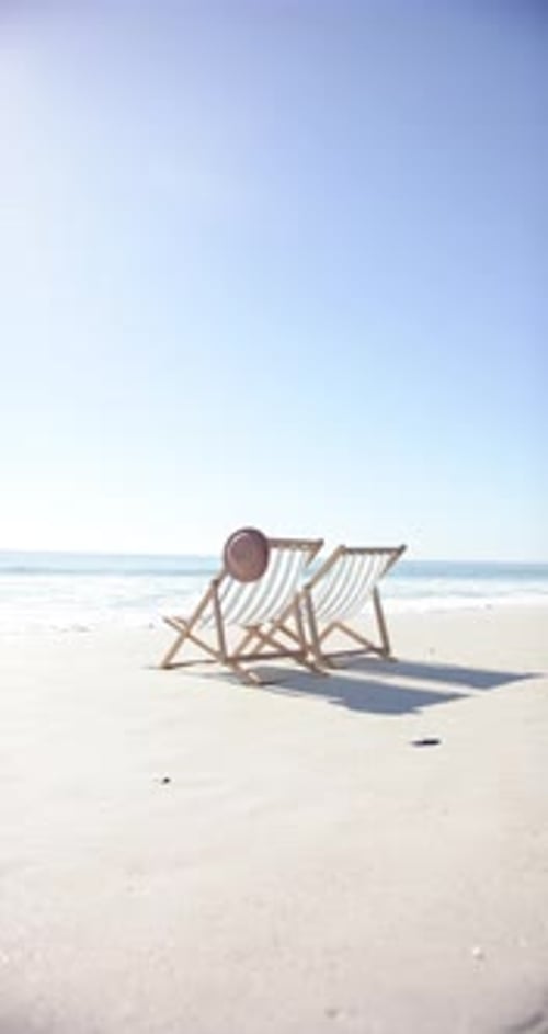 Vertical video: Two beach chairs with hat on sandy shore, relaxing by ocean