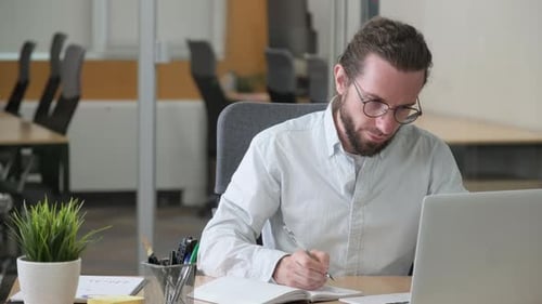 Young Adult Man Writes in Notebook at Office Desk