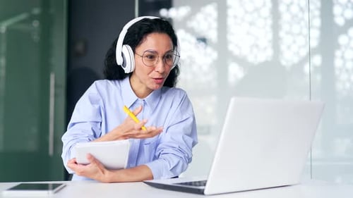 Female employee in headphones listens to online e-learning, video course sitting in office. Woman