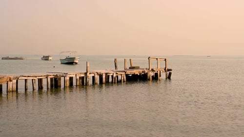 Alter Holzpier auf ruhigem Meerwasser über dem Sonnenuntergangshimmel mit Booten im Hintergrund, Bahrain.