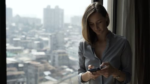 Woman using smartphone near window with city view