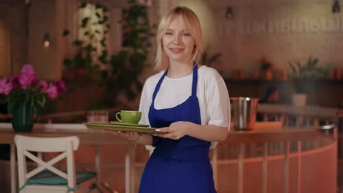 Ein blondes Mädchen in einer blauen Schürze hält ein Tablett mit einer grünen Untertasse und einer Tasse in den Händen und schaut
