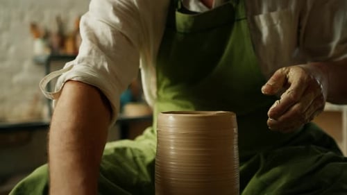 Close-up of potter's hands covered with clay making beautiful vase on throwing wheel in pottery