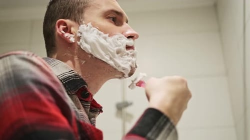 Homem se barbeando no espelho do banheiro enquanto se prepara para o dia seguinte com creme de barbear no rosto