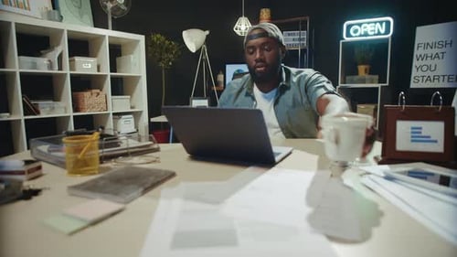 Focused african american businessman drinking coffee and typing at night in modern office