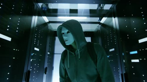 Masked Hacker in a Hoodie Walks Through Corporate Data Center with Rows of Working Rack Servers.