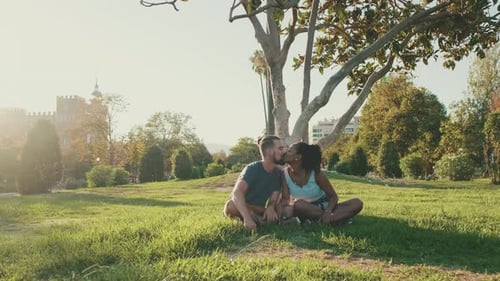 Happy smiling interracial couple talking while sitting on the grass in the park. Backlight