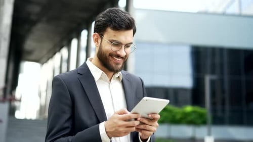 Smiling businessman is using digital tablet while standing on street near office building.