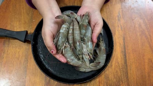 Hands Holding Raw Shrimp Over Skillet on Table