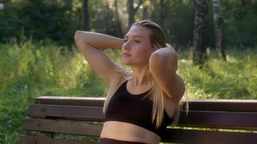 Attractive Young Woman Straightens Her Hair While Sitting on Bench in Park or Forest
