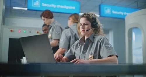 Airport Security Checkpoint Female TSA Officer Wearing Headset Works on Laptop