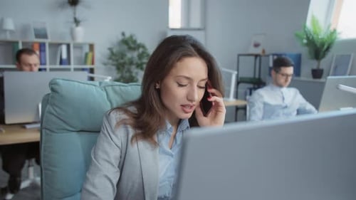 Beautiful Young Businesswoman Speaks on Phone Working on Laptop Computer in Office On Background
