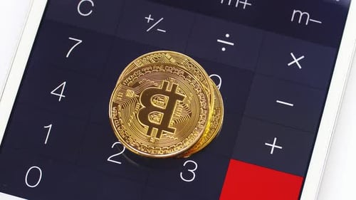 Man legt goldene Bitcoins eins nach dem anderen auf einen Taschenrechner auf einem Smartphone