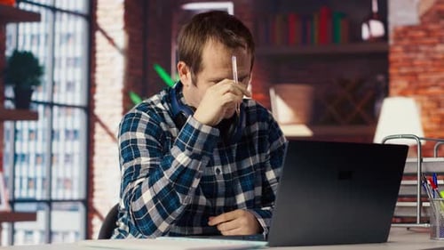 Stressed Man in Home Office Scratching Head with Pen Feeling Overworked