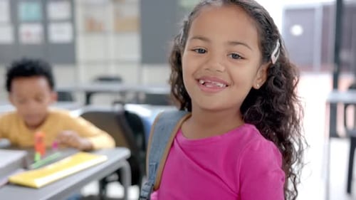 In school, smiling girl with backpack standing in classroom, looking at camera