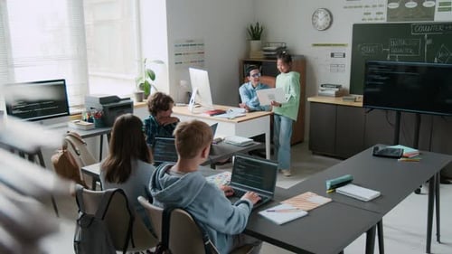 Teacher Checking Assignment of Girl, While Other Students Working on Laptops