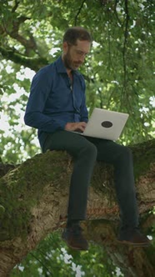 Remote Work in Nature A Man Sitting Comfortably on a Tree Branch with a Laptop Computer