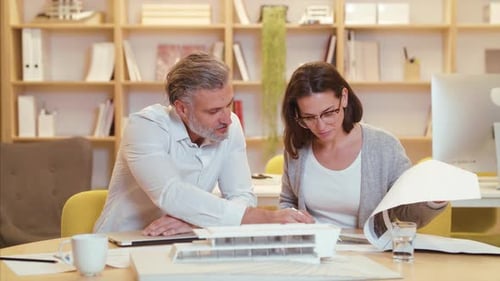 Architects With Model Of House Sitting At The Desk Indoors In Office, Working.