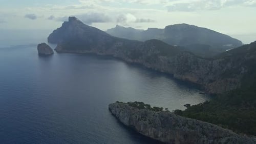 An aerial view captures a dramatic rocky coastline jutting into a calm, deep blue sea. A solitary sa