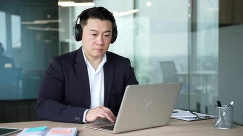 A focused asian businessman in wireless headphones works on a laptop sitting at a workplace