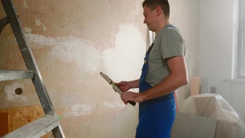 Man in Blue Construction Overalls Takes Water Level and Makes Marks with Yellow Pencil on Brown Wall