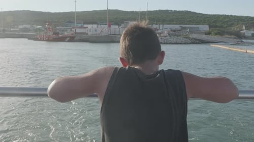 boy leaning on the stern of a boat moving away from the port