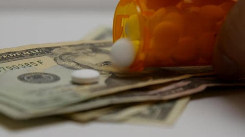 Pill bottle pours out white pills onto stacks of money.