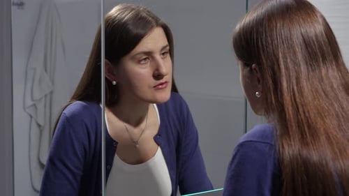 Sad young brunette woman looking at herself in mirror