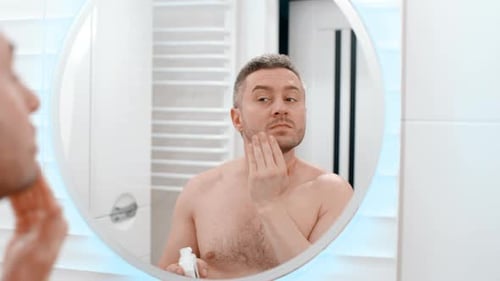 Man Applies Lotion to Face in Bathroom Mirror
