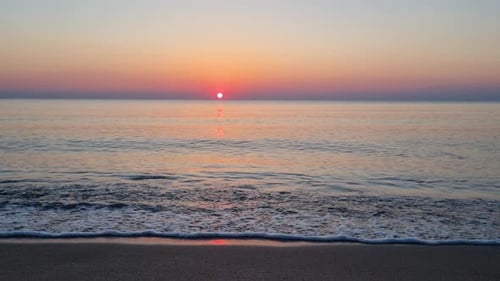 Calm water surface and sea waves splash on sandy beach at sunrise. Slow motion