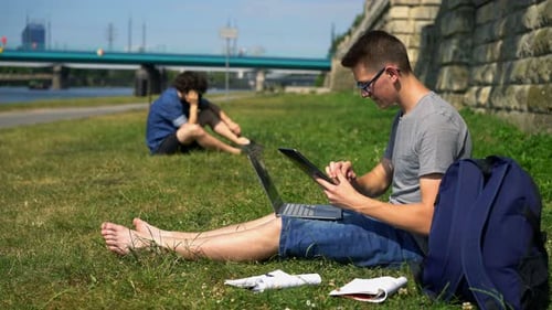 Teenage boy with tablet and laptop studies online in a city park by the river