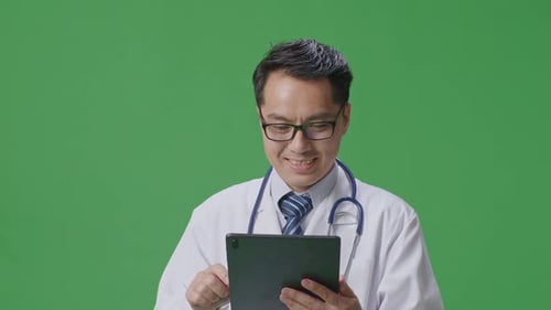 Asian Male Doctor With Stethoscope Using A Tablet While Standing On Green Screen Background