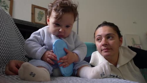 Baby sitzt auf der Couch und kaut auf blauem Objekt, Mutter schaut liebevoll zu, spielerischer Moment drinnen, Baby