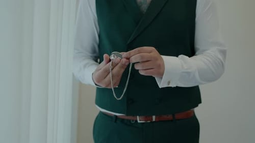 Groom adjusting a pocket watch chain, dressed in a dark green vest and white dress shirt, preparing