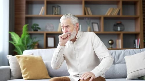 Elderly senior gray haired man coughing while sitting on sofa in living room at home. Sick mature