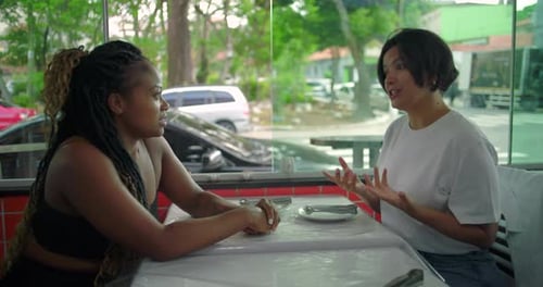 Two diverse women engaged in animated conversation at restaurant table, smiling and enjoying casual