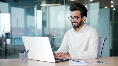 A young businessman in a white shirt typing on a laptop while sitting at a workplace in office.