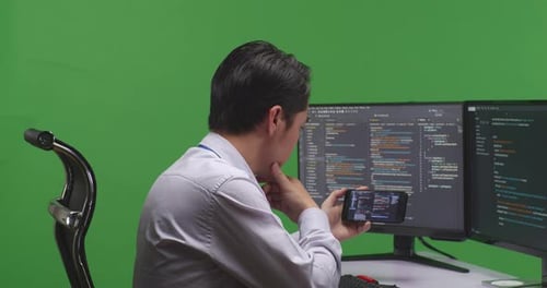 Developer Working With Mobile Phone While Write Code With Multiple Computer Screens In Green Screen