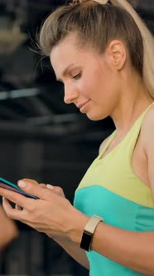 Vertical of Blonde Woman Walking on a Treadmill in the Gym and is Typing a Message on Phone