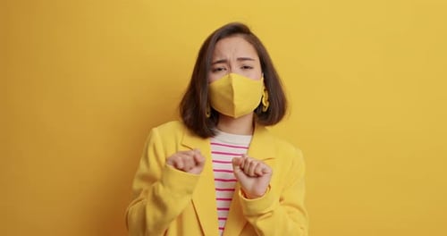 Sorglose asiatische Frau tanzt mit erhobenen Armen und trägt Gesichtsmaske auf gelbem Hintergrund