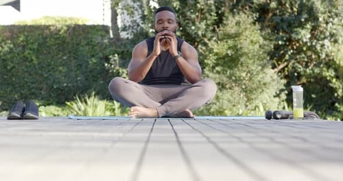 Focused african american man doing yoga meditation on deck in sunny garden, slow motion