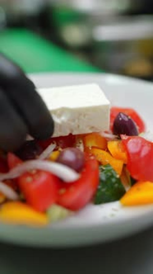 Gloved Hands Placing a Block of Feta Cheese on a Colorful Greek Salad with Tomatoes Cucumbers Onions
