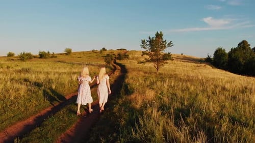 Two Young Blonde Women in Summer Light Dresses Holding Hands and Walking on the Field at Warm