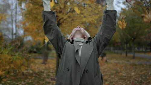 Cheerful blonde woman enjoys autumn day throwing yellow leaves in the park