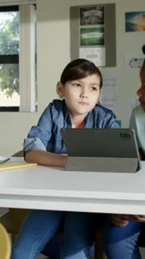 Vertical video: Glancing child wearing denim shifting gaze right to peer and viewing tablet at desk